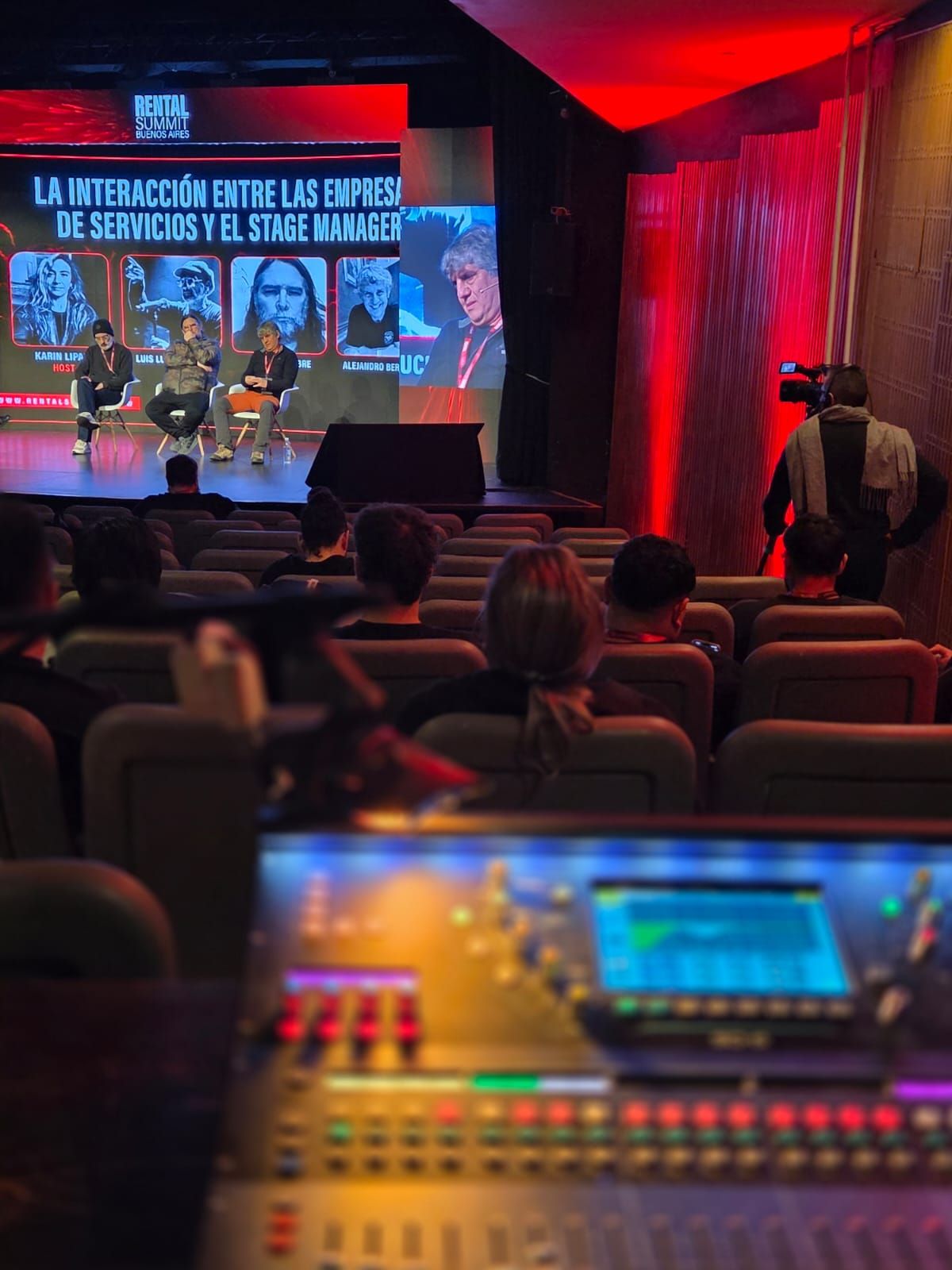 Consola de audio en primer plano y el público del Summit en la sala iluminada de rojo