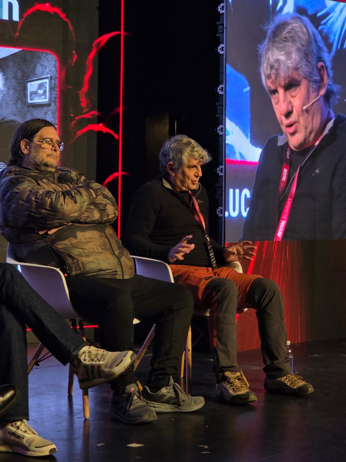 Dos panelistas conversando en el escenario con sus retratos en la pantalla LED